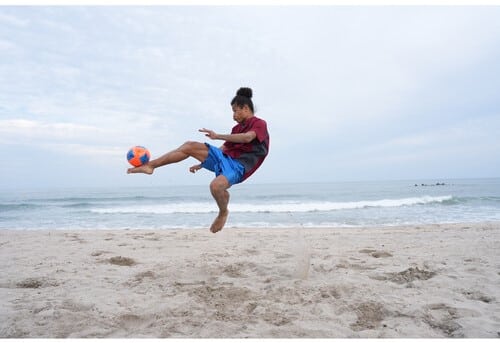 Un homme joue au foot sur la plage avec un ballon orange et bleu, capturé avec un Sony A7 V.