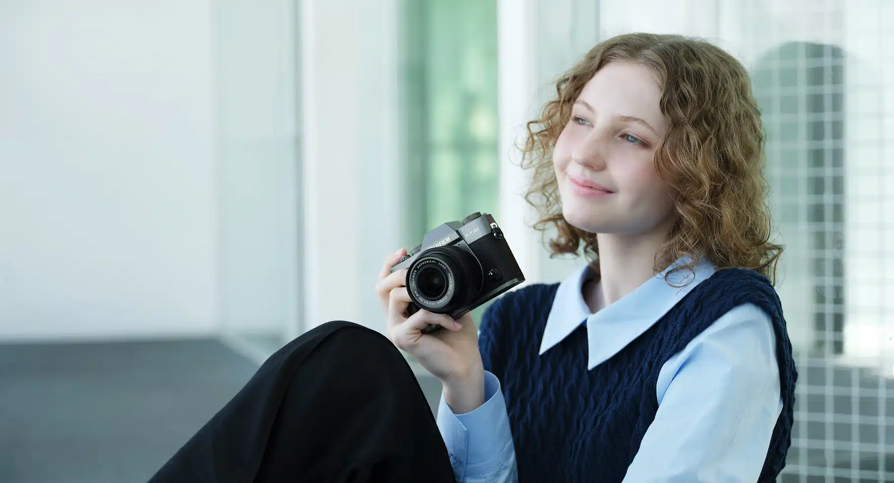 Fujifilm X-T30 III&nbsp;: Jeune femme tenant un appareil photo compact Fujifilm X-T30 III, regard rêveur.