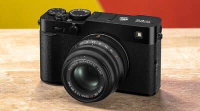 Fujifilm X-E5 noire de côté avec objectif Fujinon 35mm sur une table en bois, fond jaune et rouge.