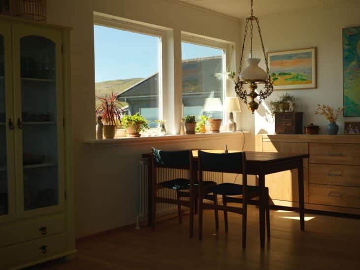 Intérieur ensoleillé avec table à manger, chaises, buffet en bois et plantes près de la fenêtre. Éclairage ambiant.