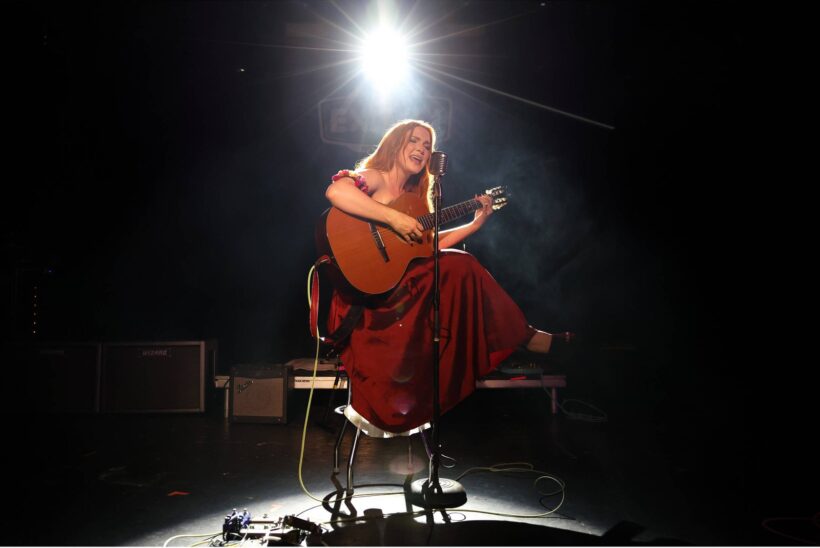 Guitariste en robe rouge assise sur une scène sombre, éclairée par un projecteur