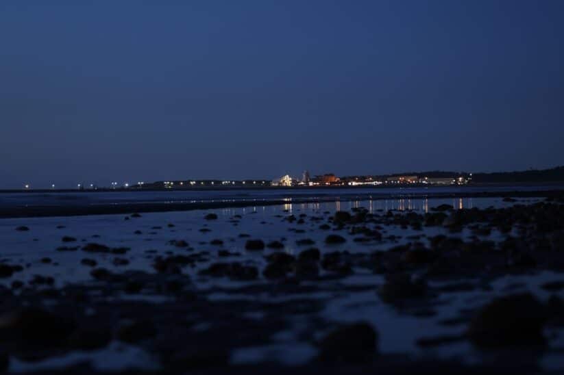 Bord de mer à l'heure bleue, rochers au premier plan et lumières d'une ville à l'horizon