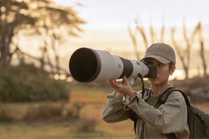 Photographe en casquette visant avec un Sony A7R VI monté sur un long téléobjectif blanc, dans la savane au lever du jour