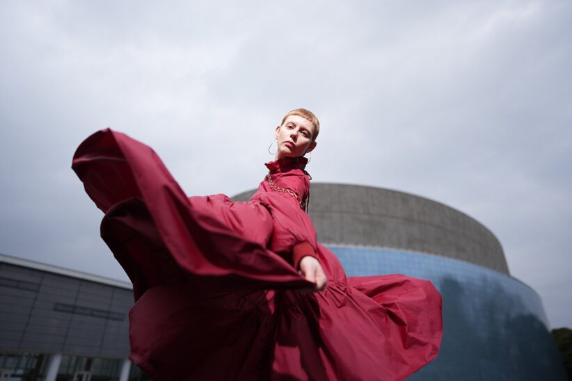 Danseuse en robe rouge ample saisie en plein mouvement devant un bâtiment arrondi, sous un ciel gris