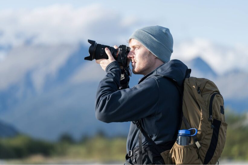 Randonneur en bonnet cadrant avec un Sony A7R VI devant des montagnes enneigées